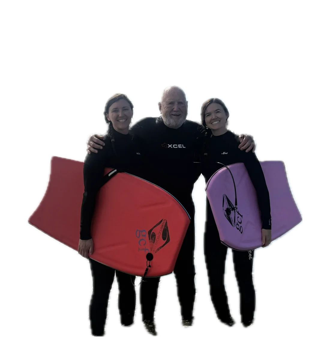 Bob standing between two women in wetsuits, each holding a bodyboard, arms around one another on the beach